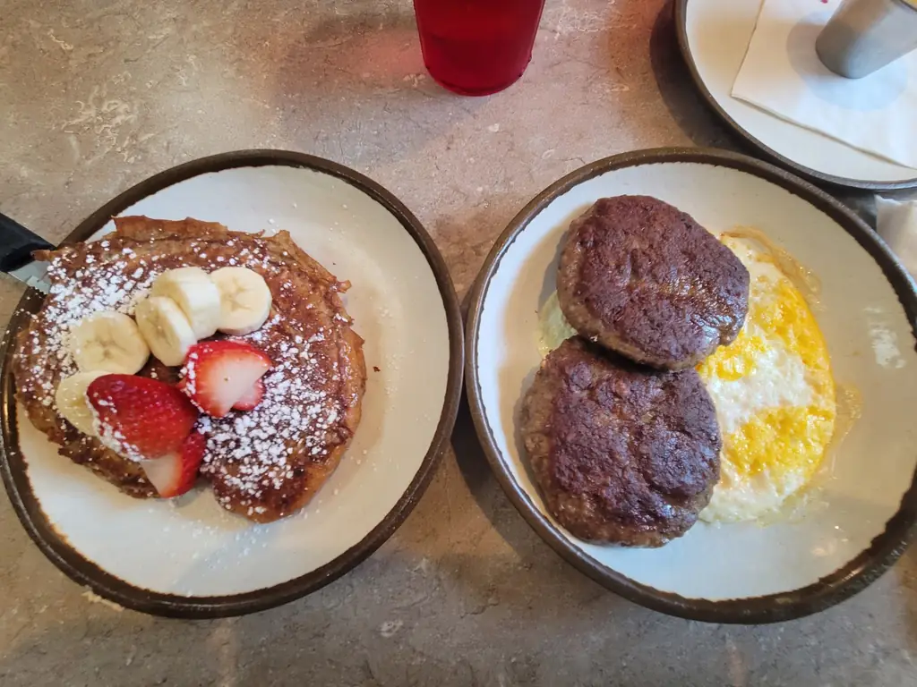 Brioche French toast with sliced strawberries, bananas and powdered sugar beside sausage patties and eggs at 43rd Street Deli